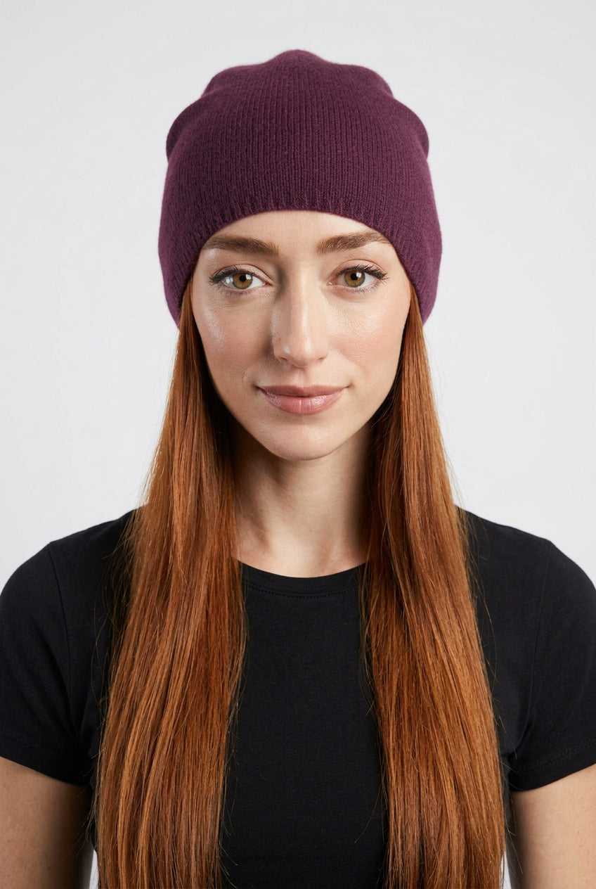 A woman with long red hair wearing a dark plum purple ribbed knit beanie hat, paired with a black top, photographed against a plain light gray background.