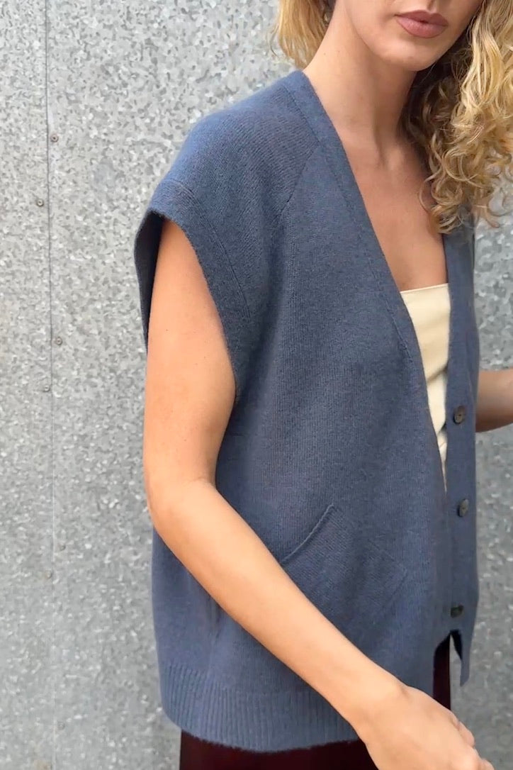 model wearing a blue cashmere sleeveless cardigan and plaid scarf against a textured wall. side view