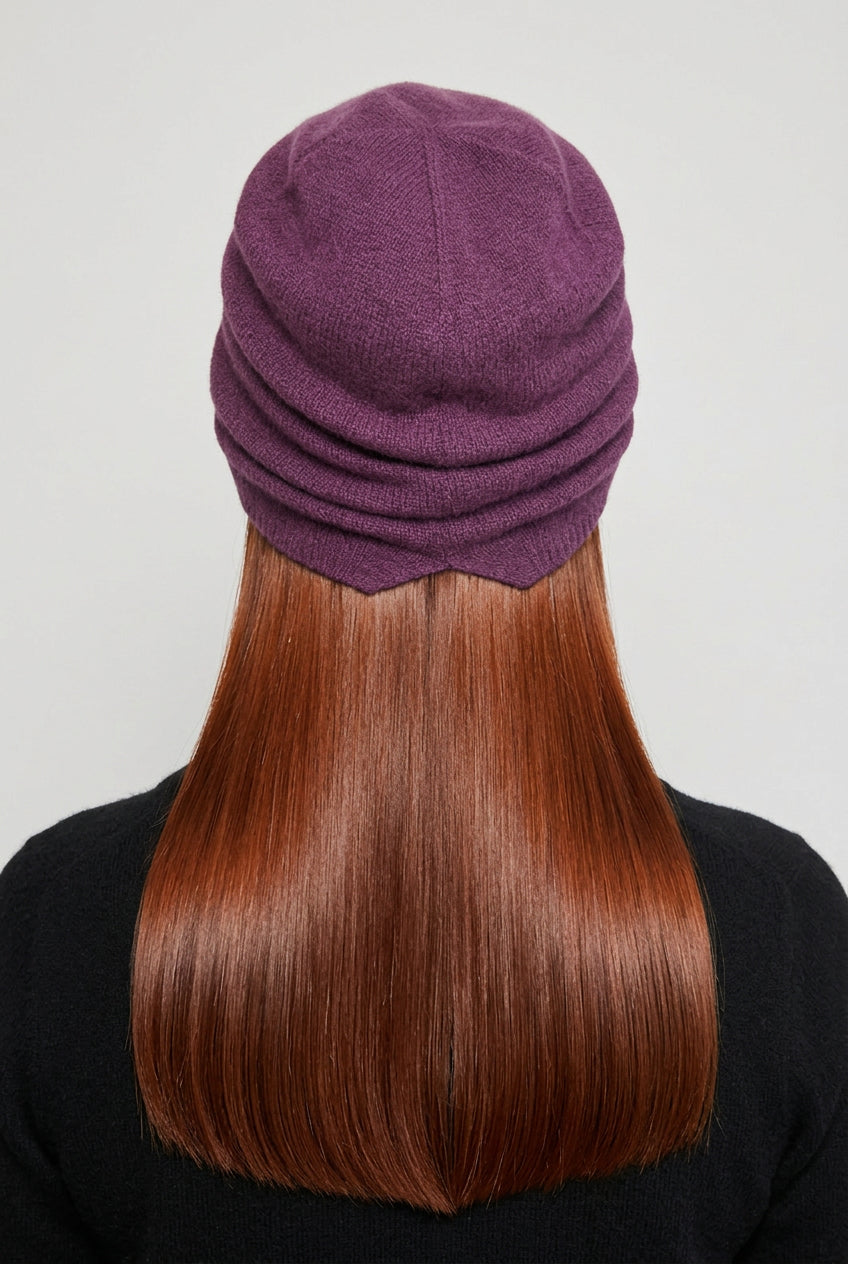 A person viewed from behind wearing a slouchy knit beanie in a muted plum purple color, with long light brown hair and a black top against a plain light background.