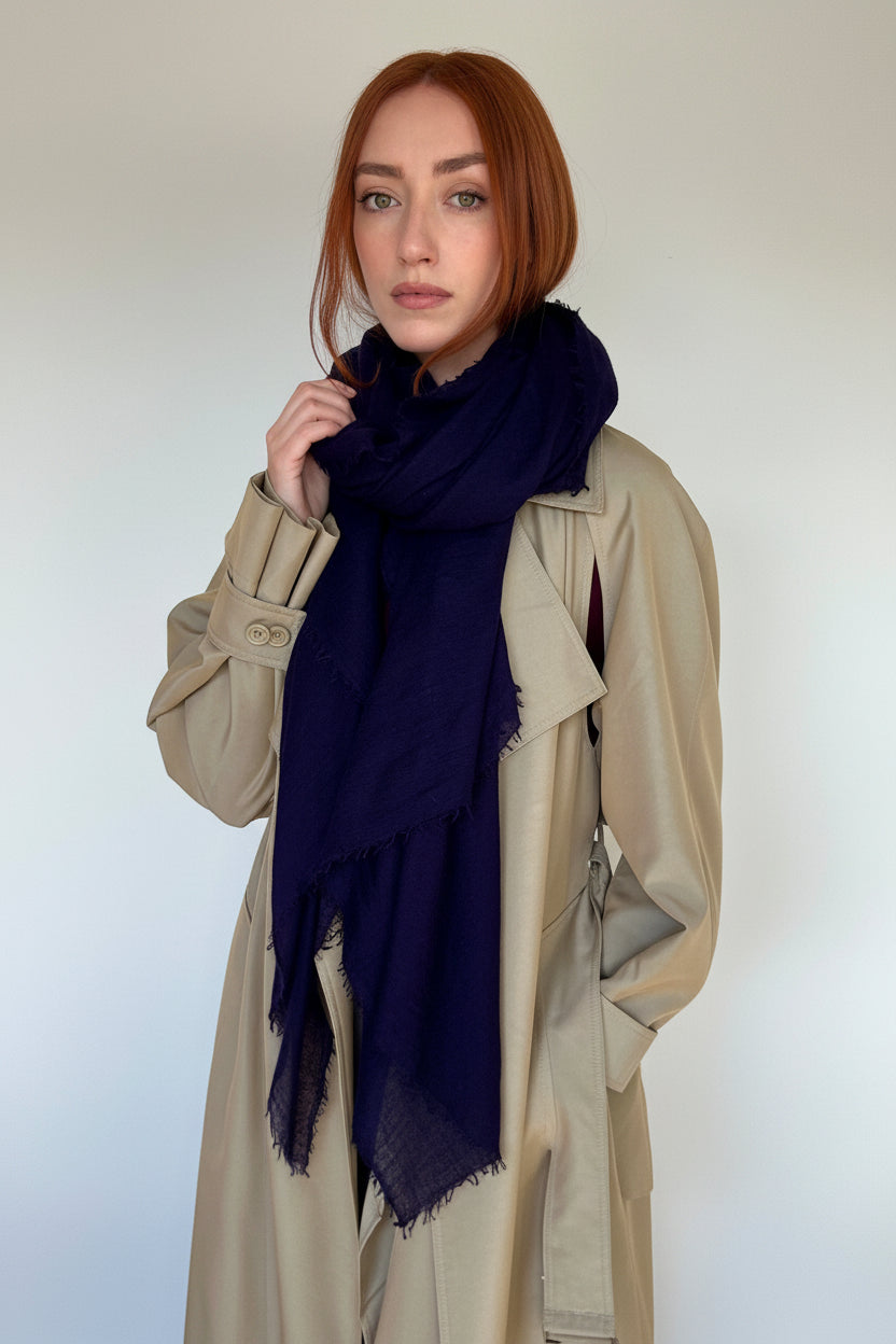 A woman with red hair wearing a beige trench coat and a dark navy blue scarf, standing against a plain light gray wall.