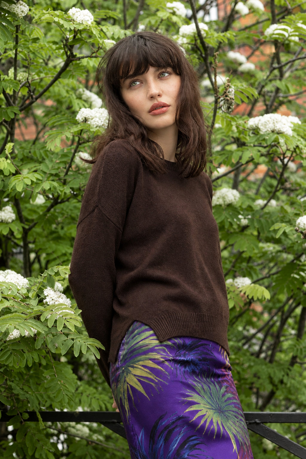 A woman wearing a dark brown knit sweater and purple. skirt posing outdoors in front of leafy green bushes with small white flowers.