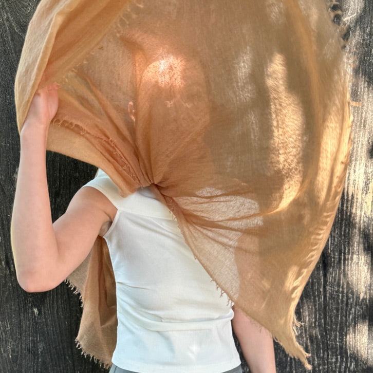 Woman with a fine cashmere scarf blowing in the wind in front of her face Marmee scarf in gold colour camel caramel