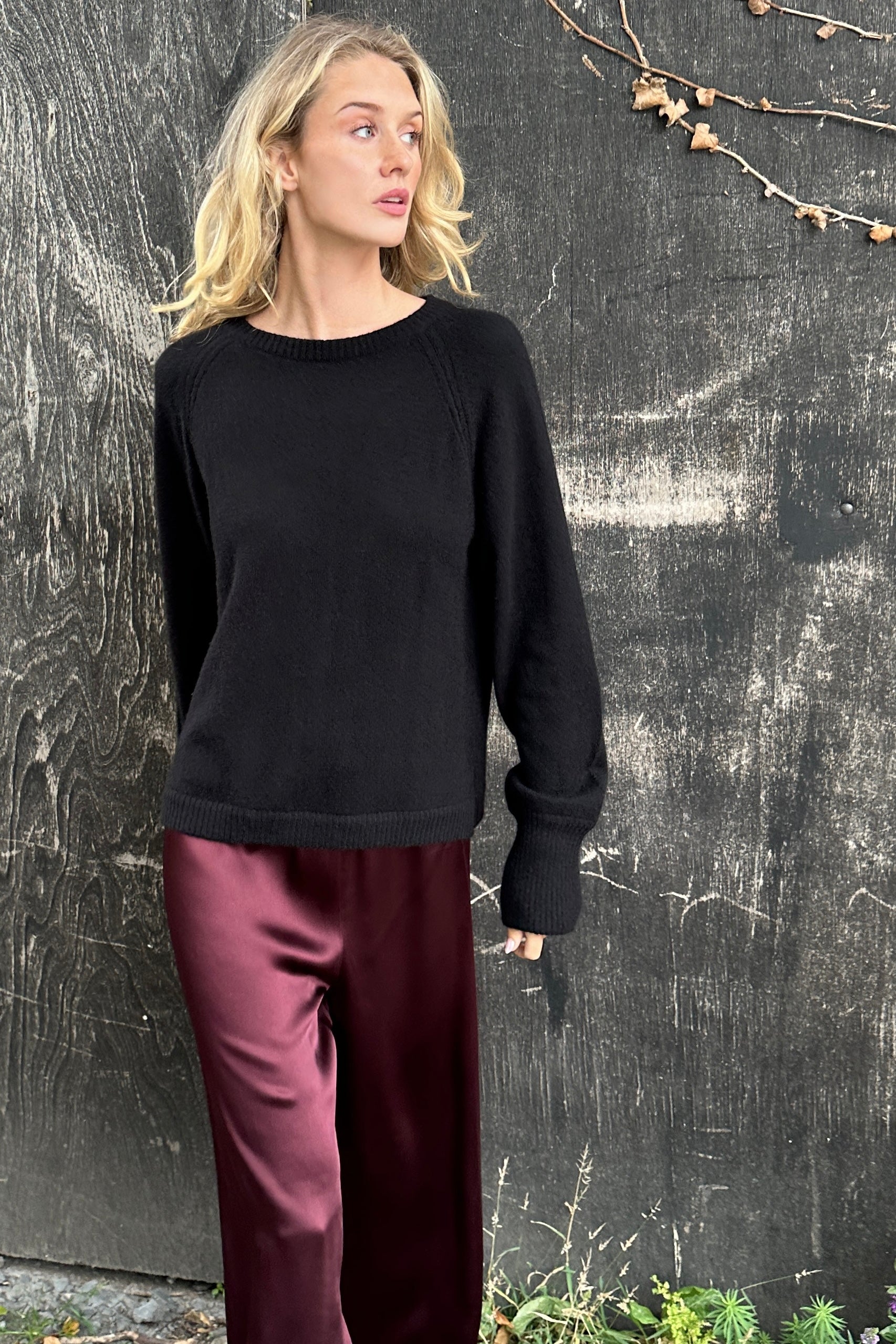 Woman wearing a black cashmere sweater and burgundy pants standing against a textured wall.