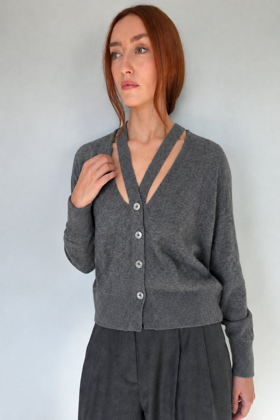 A woman with red hair wearing a dark grey melange button-up cardigan with a split V-neckline and gray trousers, standing against a plain light gray wall.