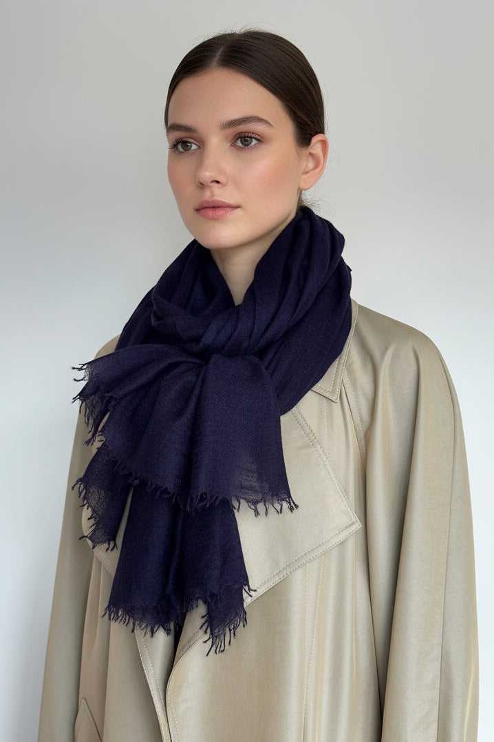 A dark navy blue scarf with fringed edges, styled on a white mannequin torso wearing a beige trench coat against a plain light gray background.