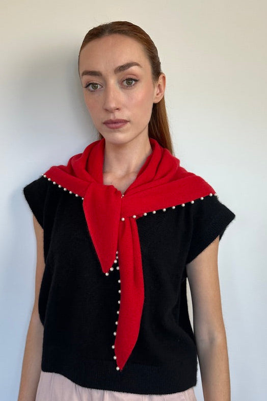A woman modeling a black knit top with a bright red scarf draped over her shoulders, decorated with small white pearl-like accents along the collar and front tie, against a plain light gray wall.
