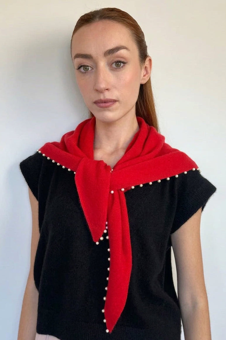 A woman modeling a black knit top with a bright red scarf draped over her shoulders, decorated with small white pearl-like accents along the collar and front tie, against a plain light gray wall.
