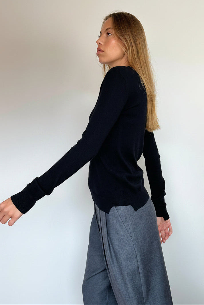 A woman wearing a black long-sleeve knit top and dark gray wide-leg trousers, standing in profile against a plain off-white wall.