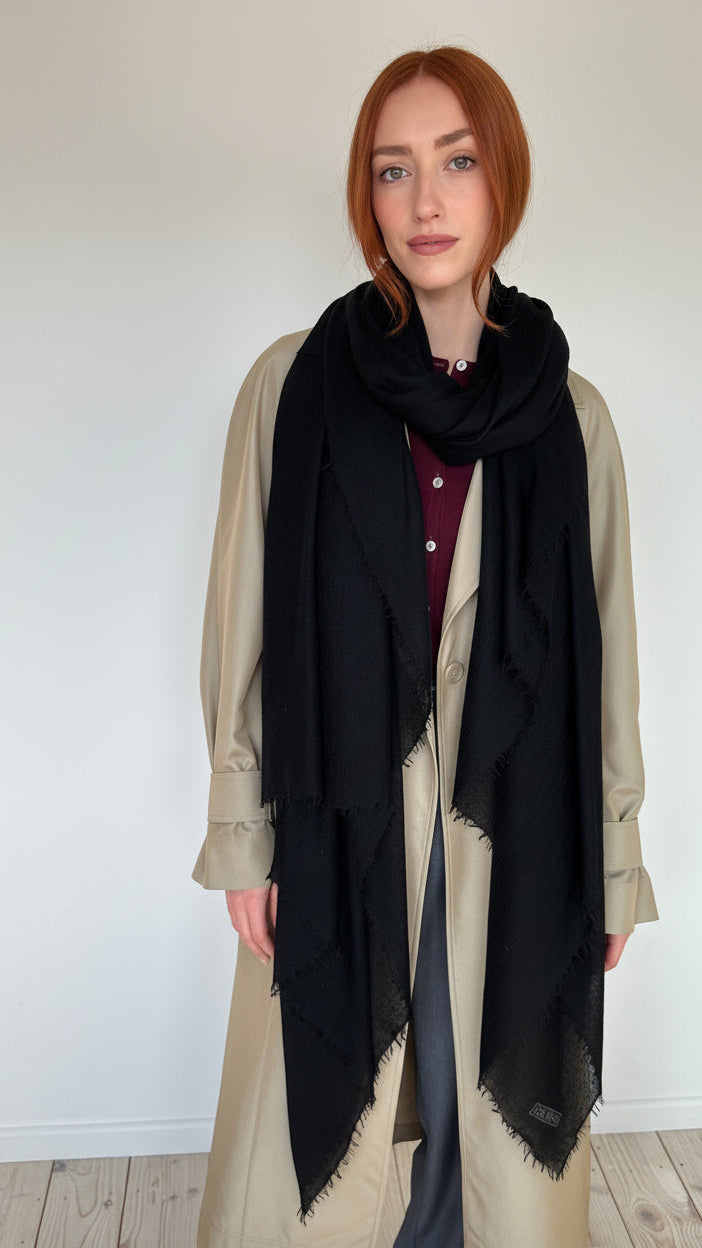 A woman with red hair wearing a beige trench coat over a burgundy top, wrapped in a long black scarf with frayed edges, standing against a plain white wall and light wood floor.