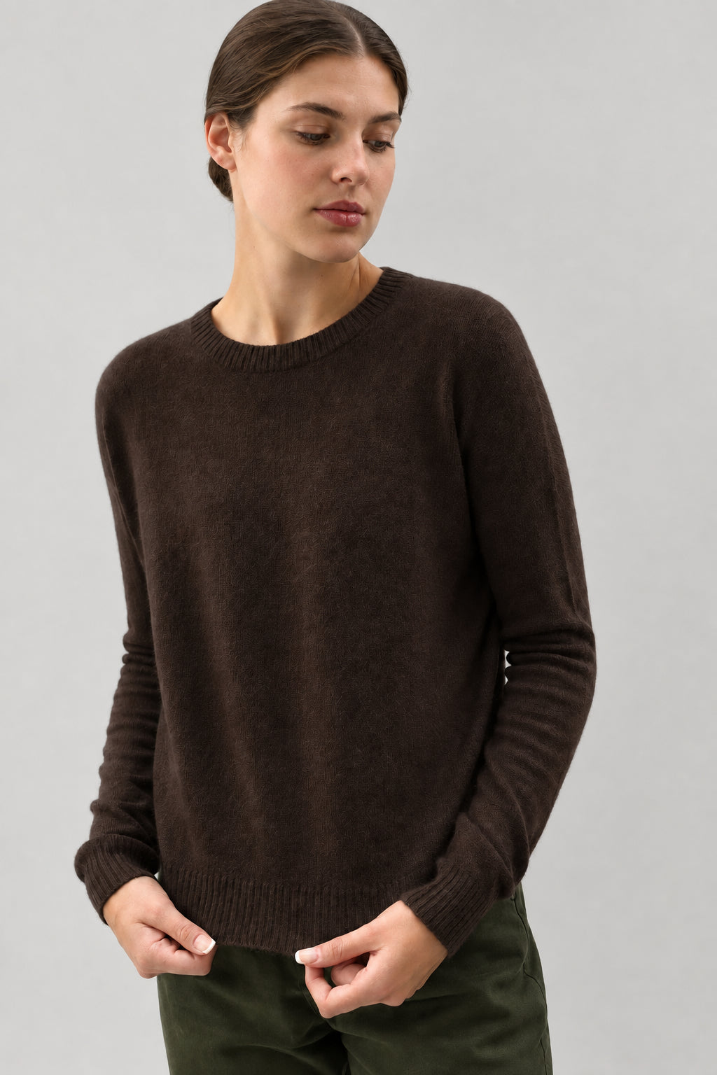 A person wearing a dark brown crewneck knit sweater, shown from the mouth down against a plain light gray background.
