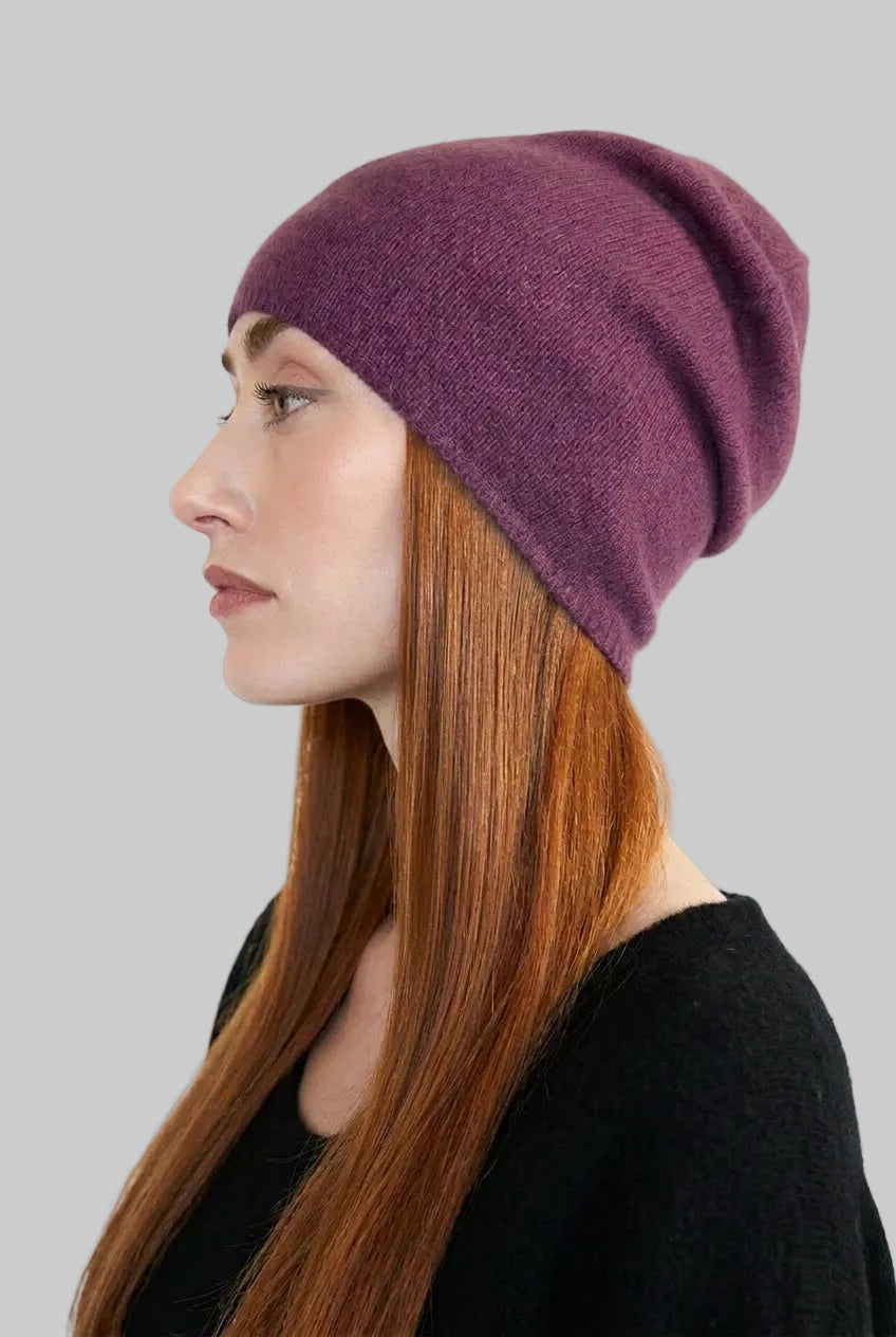 A woman in profile wearing a soft, folded knit beanie hat in a muted plum-purple color, paired with a black top against a plain light background.