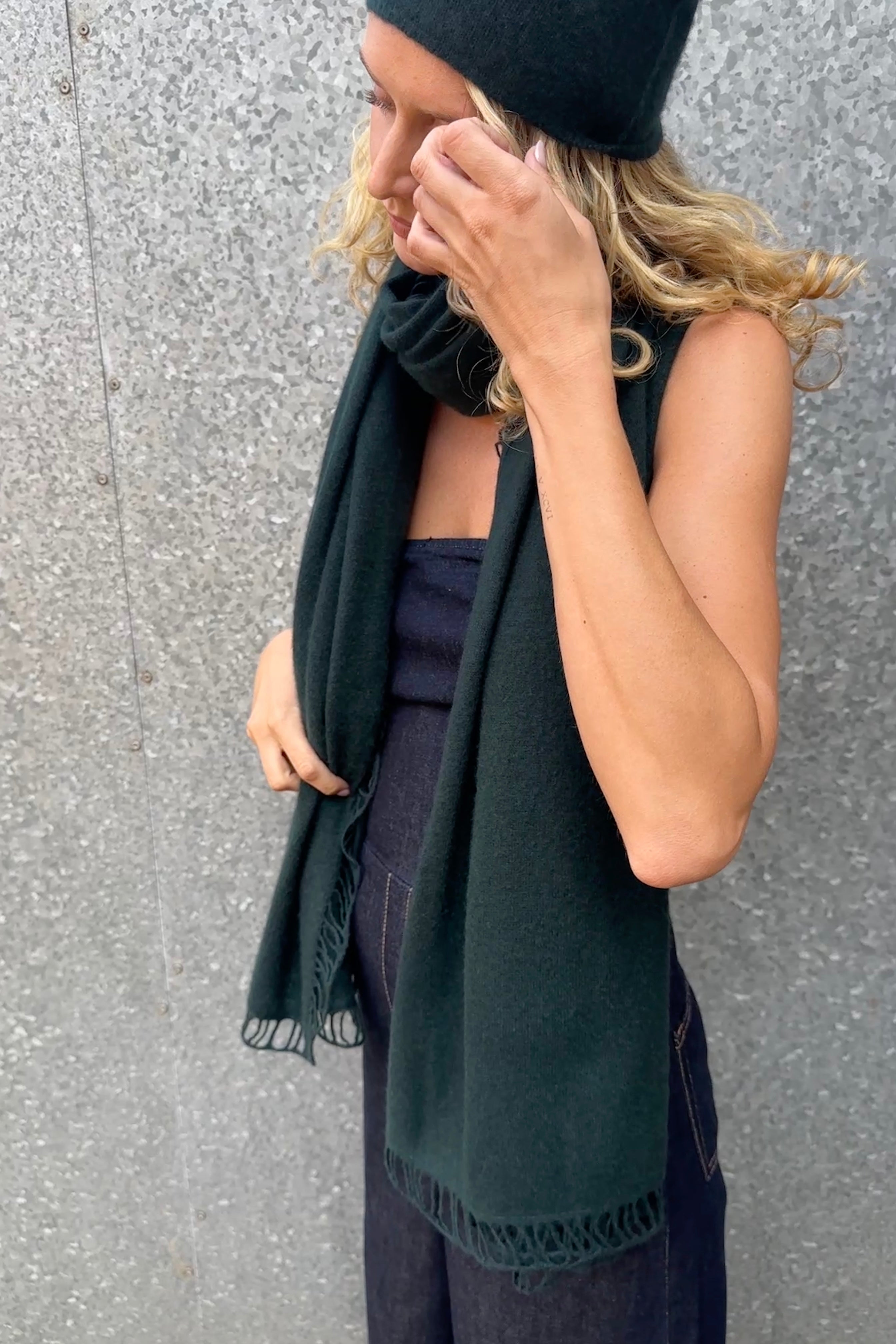 Woman wearing a dark green scarf and green beanie against a gray wall