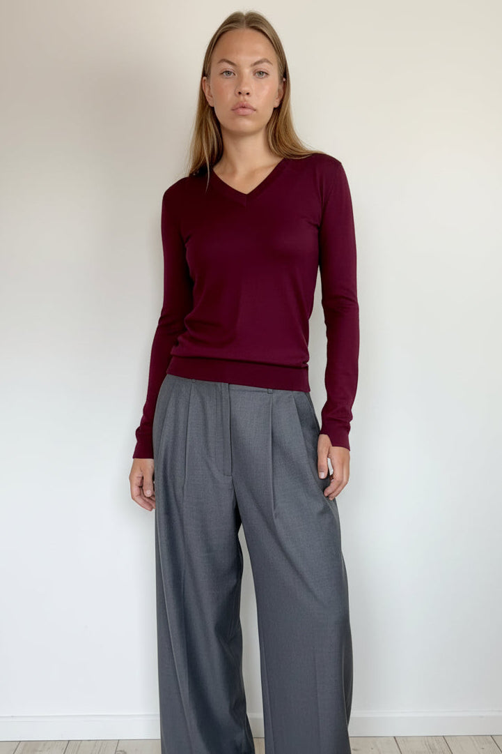 A woman modeling a long-sleeve V-neck knit sweater in deep burgundy, paired with high-waisted gray wide-leg trousers, standing against a plain white wall.