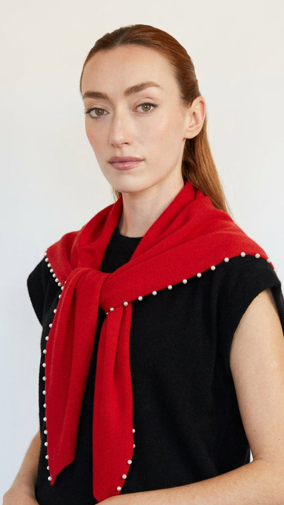 A woman with red hair tied back is shown from the side, wearing a sleeveless top with a bold red and black color-blocked design and a row of white pearls along the shoulder seam, set against a plain off-white wall.
