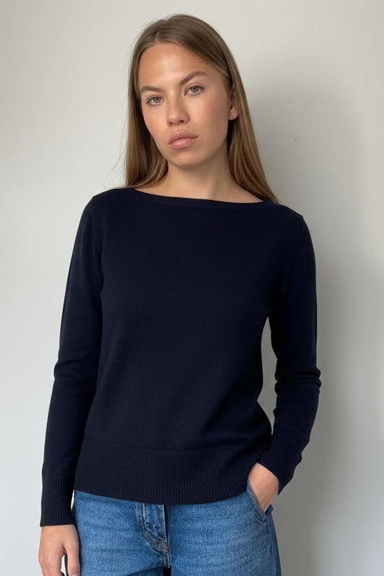 A woman wearing a dark navy long-sleeve knit top with a wide boat neckline, paired with blue jeans, standing against a plain light gray wall.