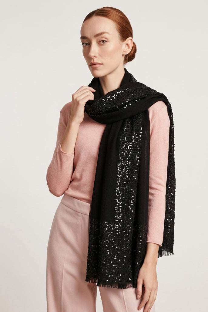 A woman modeling a black sequined scarf with fringe, worn over a pink long-sleeve top and light pink pants, standing against a plain light gray wall.