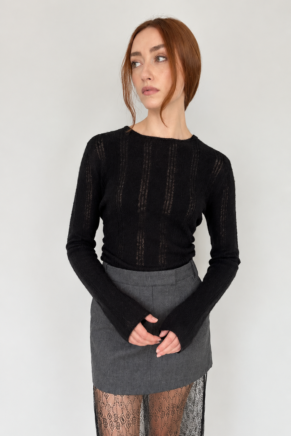 A woman modeling a black long-sleeve knit top with sheer vertical ribbing and a gray skirt featuring a sheer black lace overlay, standing against a plain light gray wall.