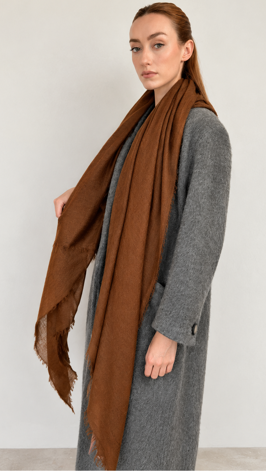 A woman wearing a long brown cashmere scarf draped over a gray coat, standing against a plain light gray wall.