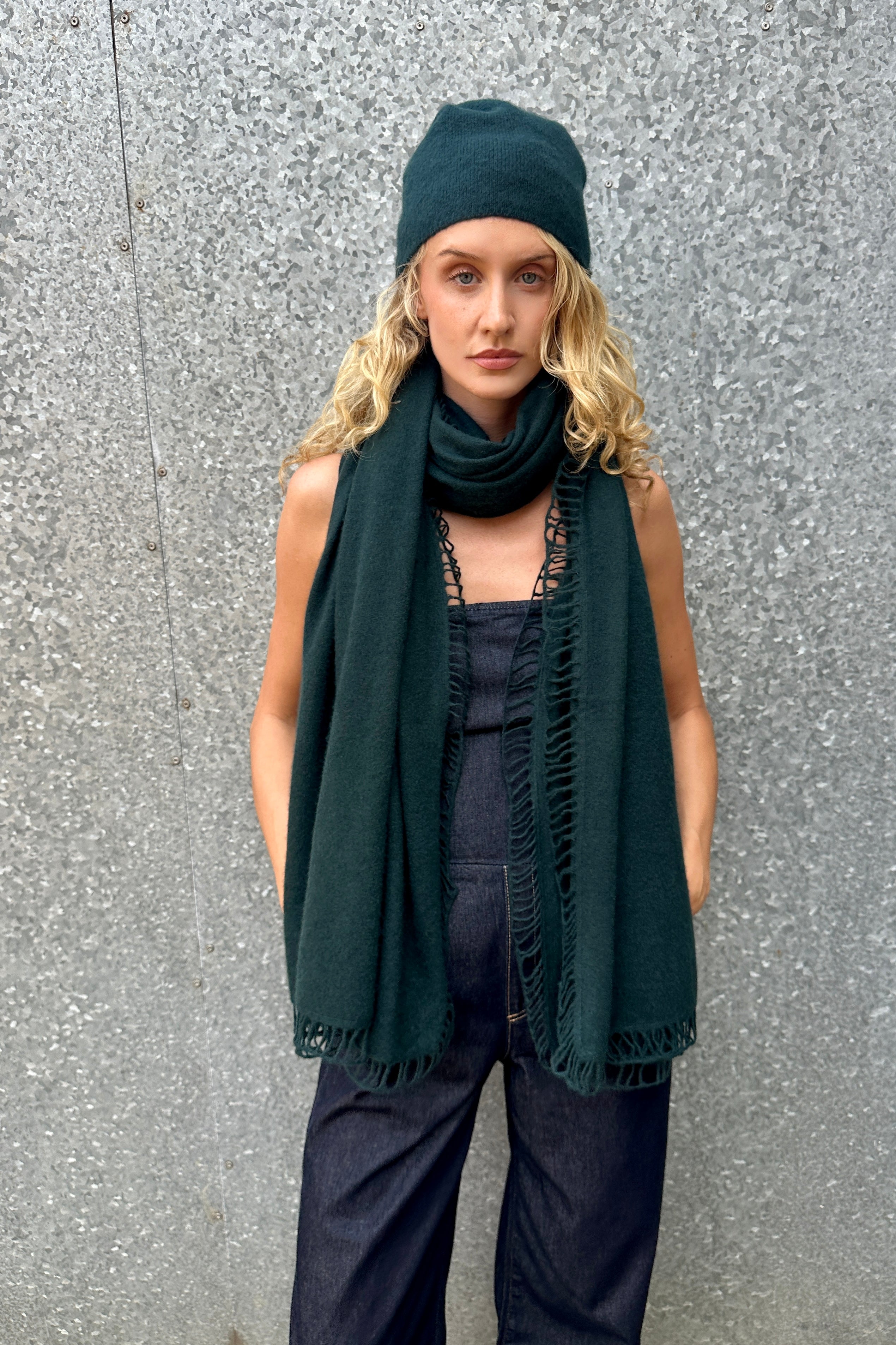 Person wearing a dark green scarf, beanie, and pants against a gray wall.