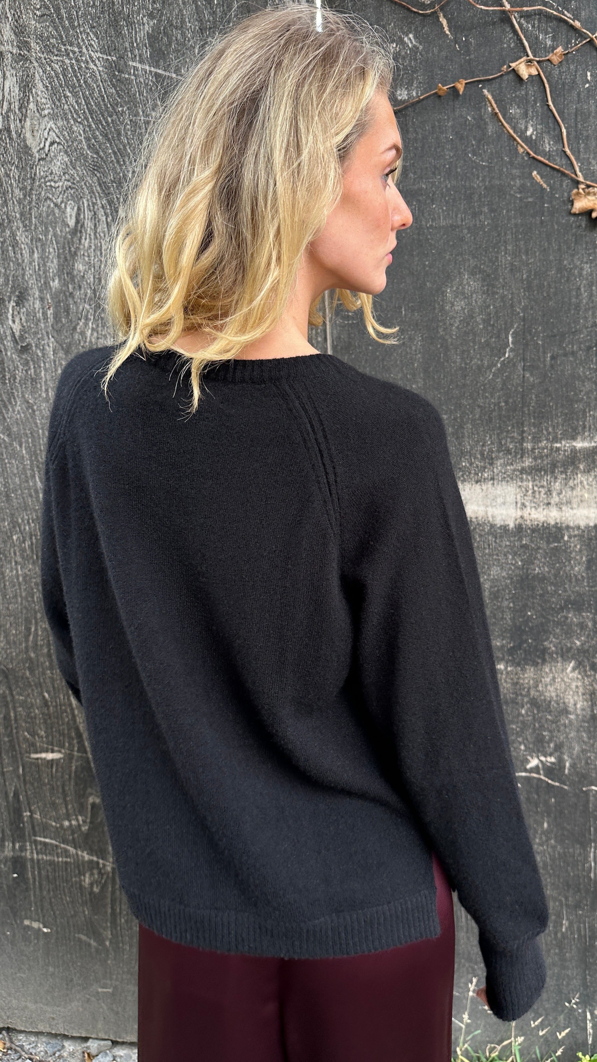 Woman wearing a black cashmere sweater and burgundy pants standing against a textured wall. back view
