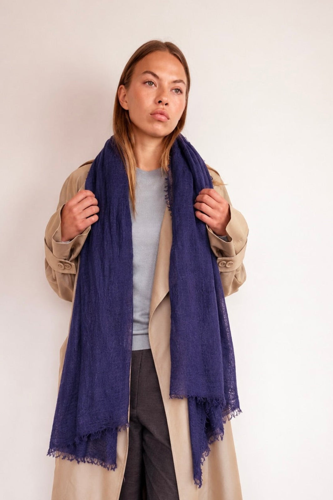 A woman modeling a cashmere navy blue fringed scarf wrapped around her neck, styled with a beige trench coat, gray top, and dark trousers against a plain white wall.