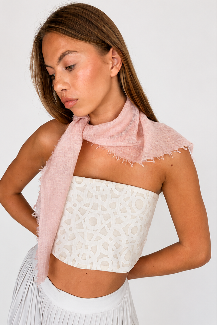 model photo showing a pale pink fringed scarf styled around the neck of a woman wearing a white strapless cropped top and white pleated pants, against a light gray studio background.