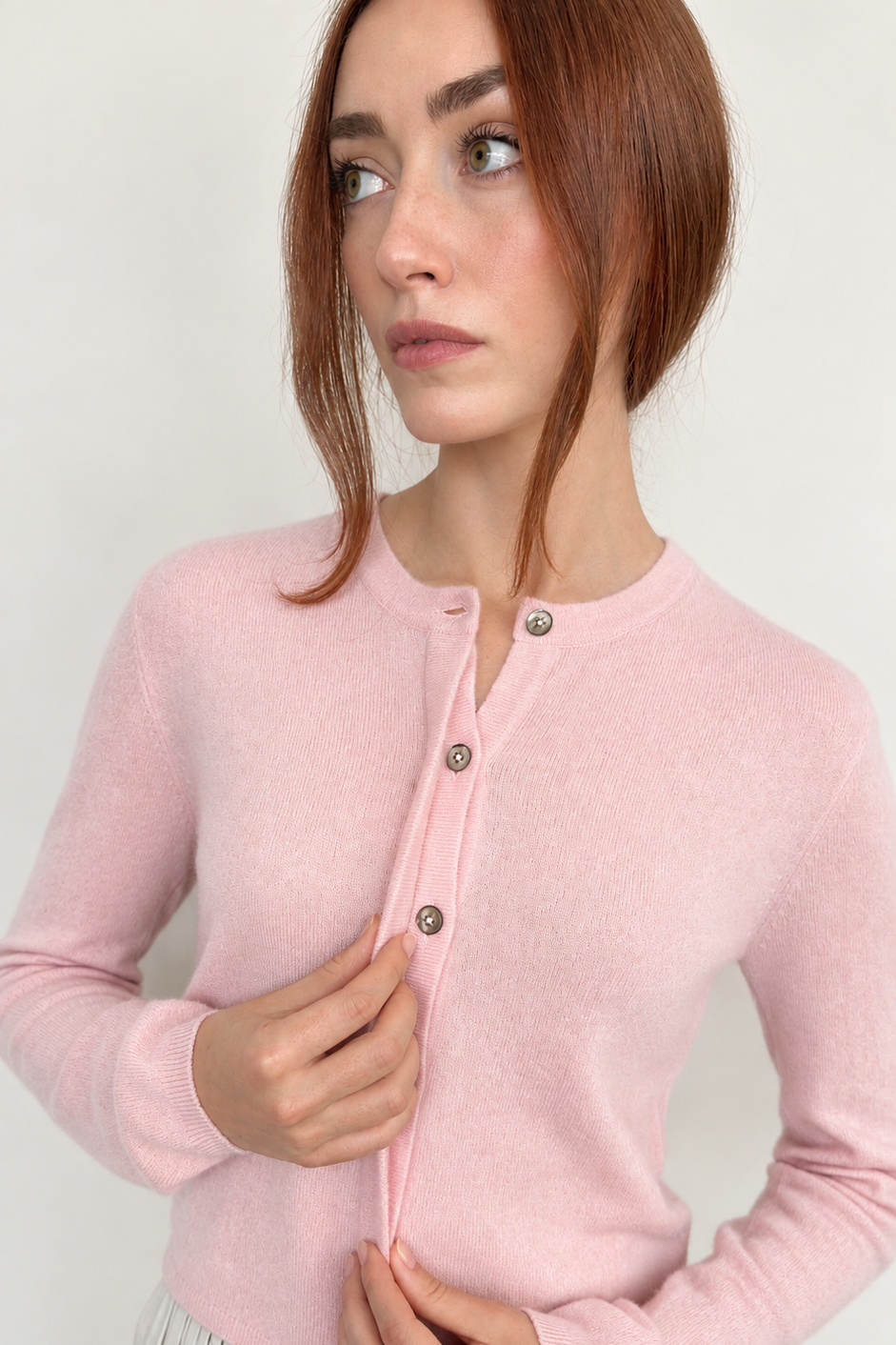 A woman wearing a light pink long-sleeve knit cardigan with a round neckline and three front buttons, paired with a white pleated skirt, against a plain light background.