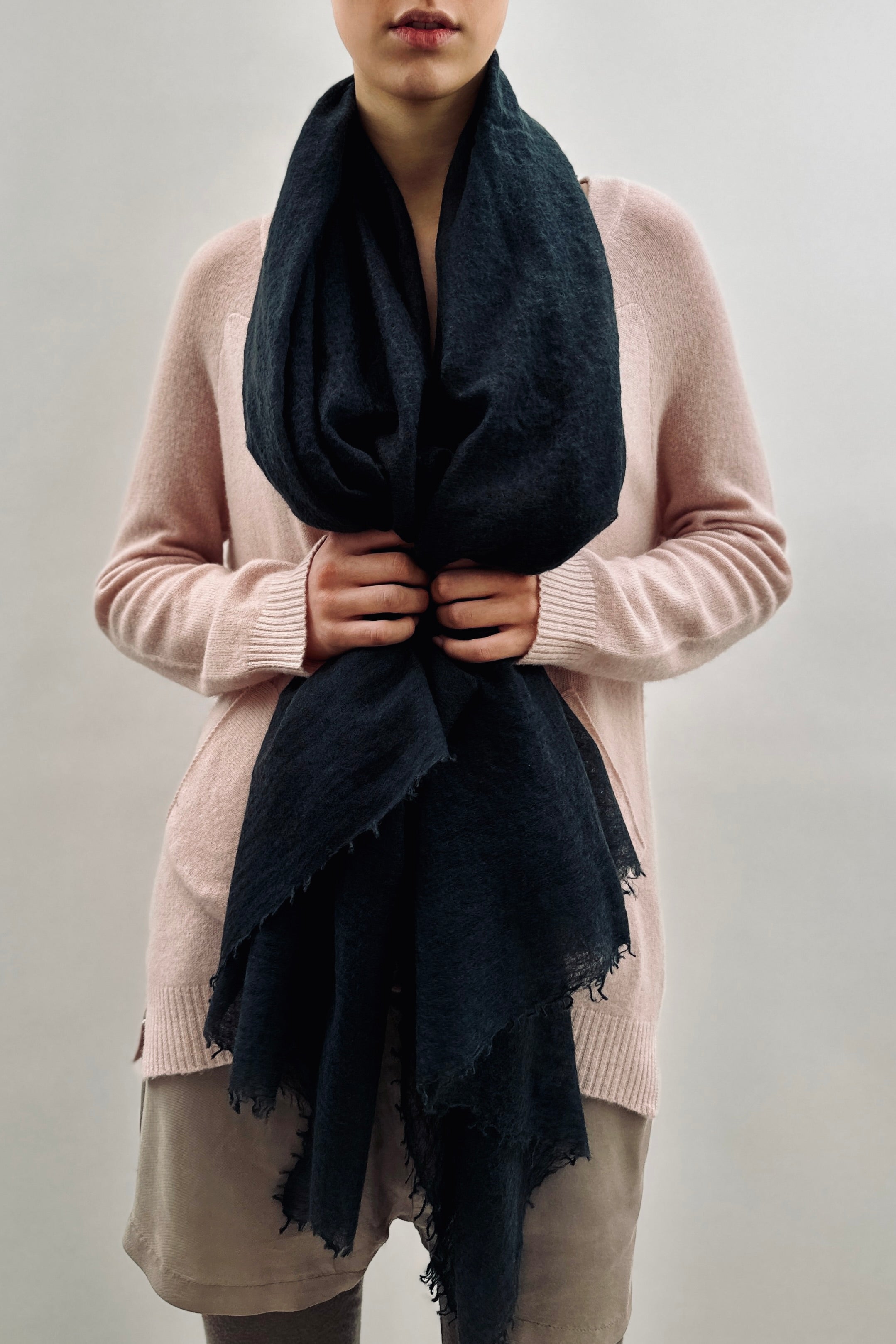 Person wearing a pink sweater and beige pants with a black scarf against a plain background