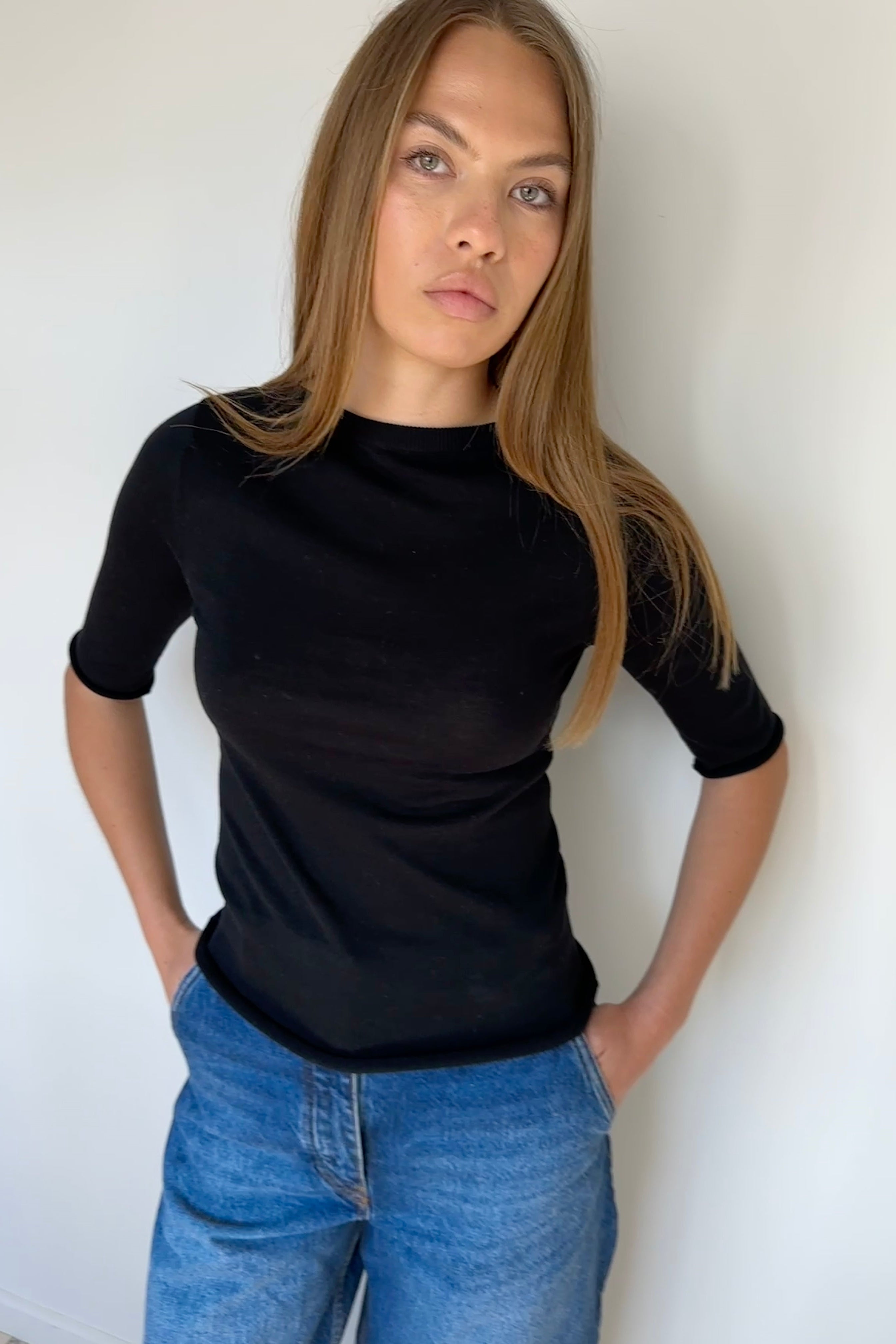Woman wearing a black top and blue jeans against a white wall