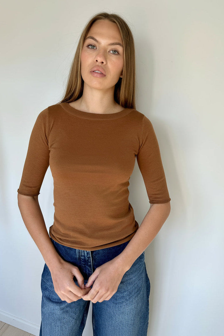 Woman wearing a brown elbow length t shirt and blue jeans against a white wall