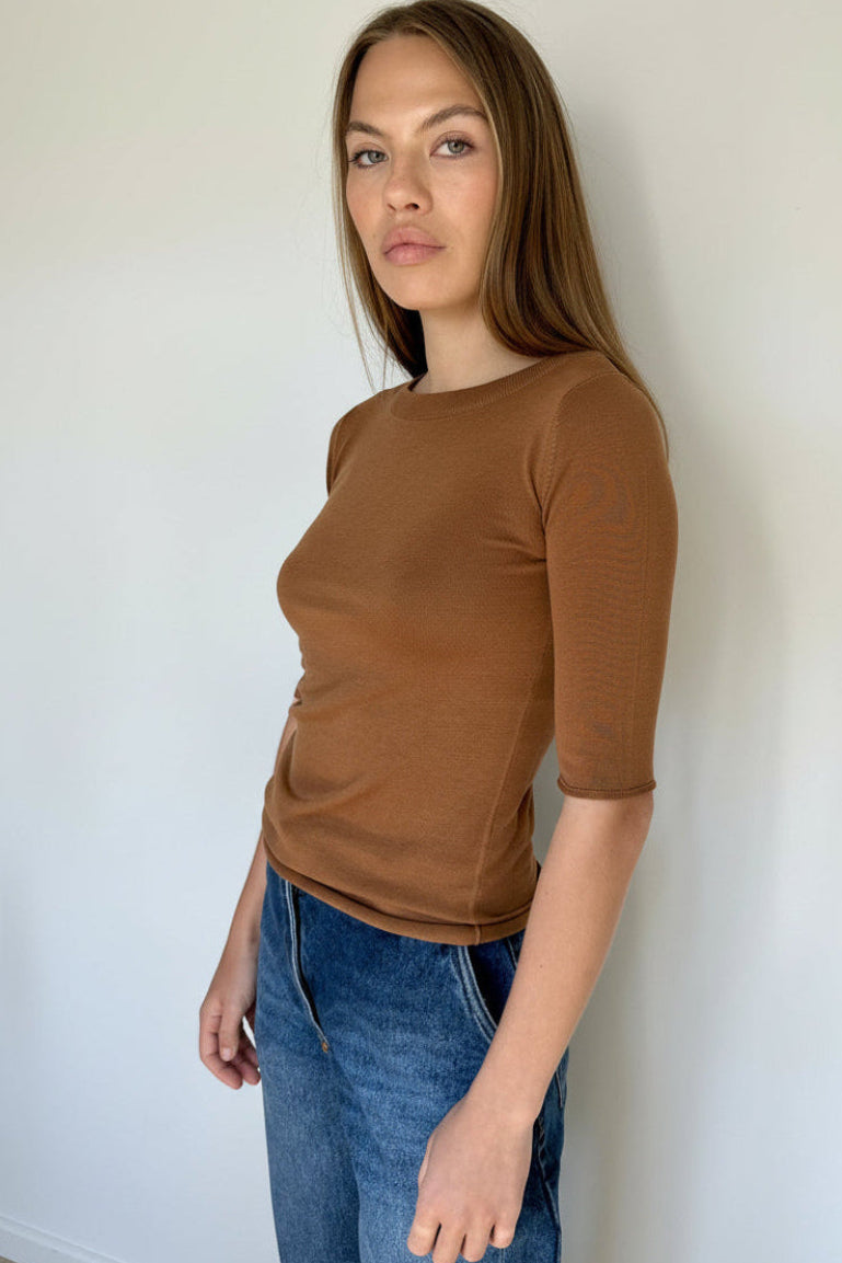 Woman wearing a brown shirt and blue jeans against a plain wall