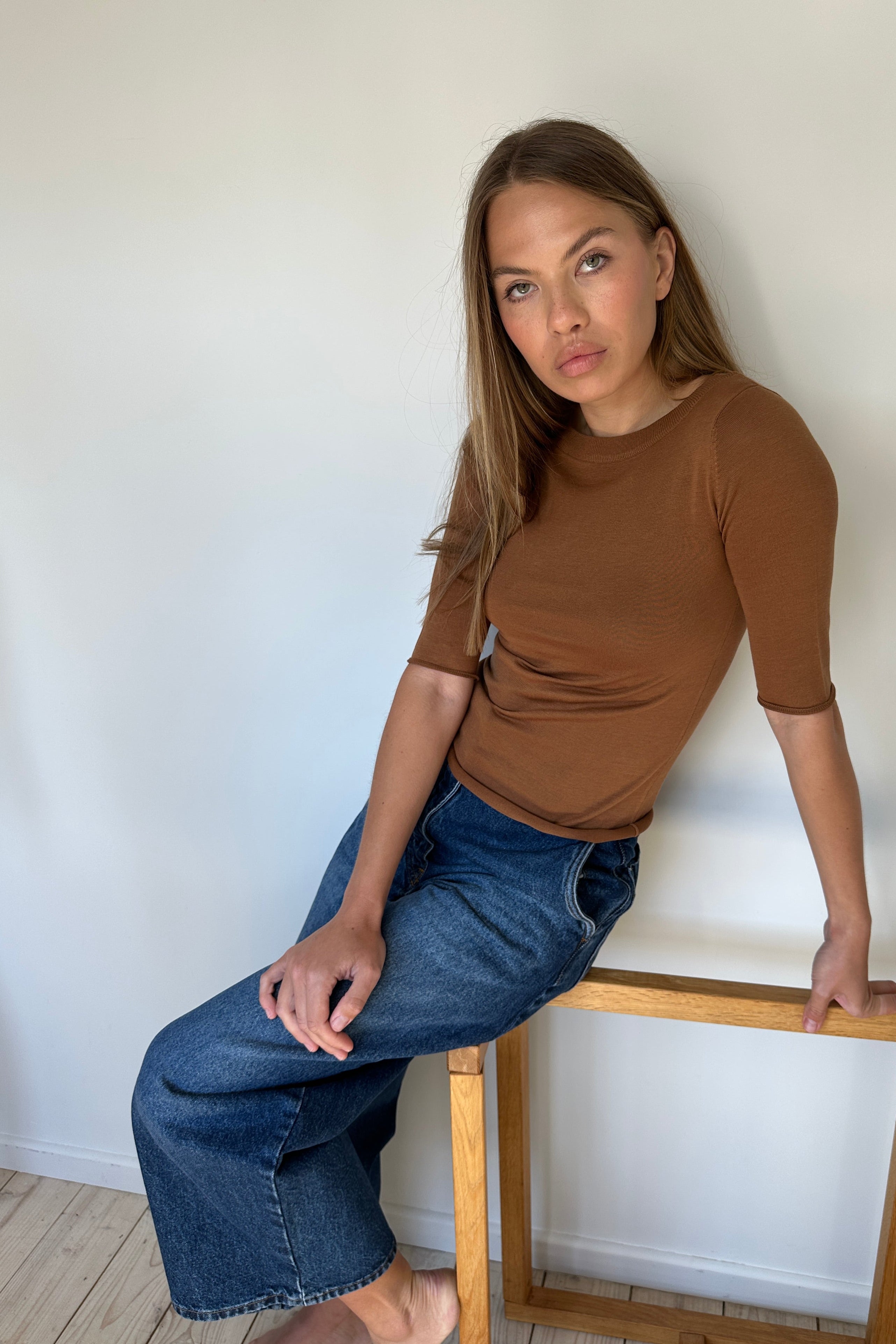 model wearing a brown top and blue jeans standing next to a wooden frame on a white floor.