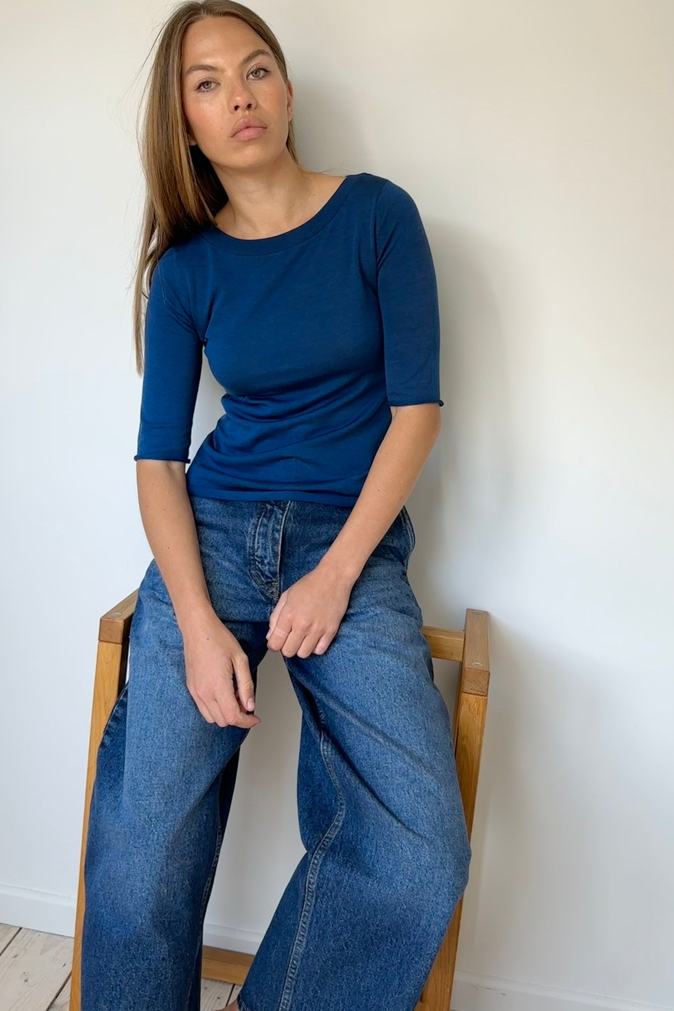 Woman wearing a blue top and jeans sitting on a wooden chair against a white wall.