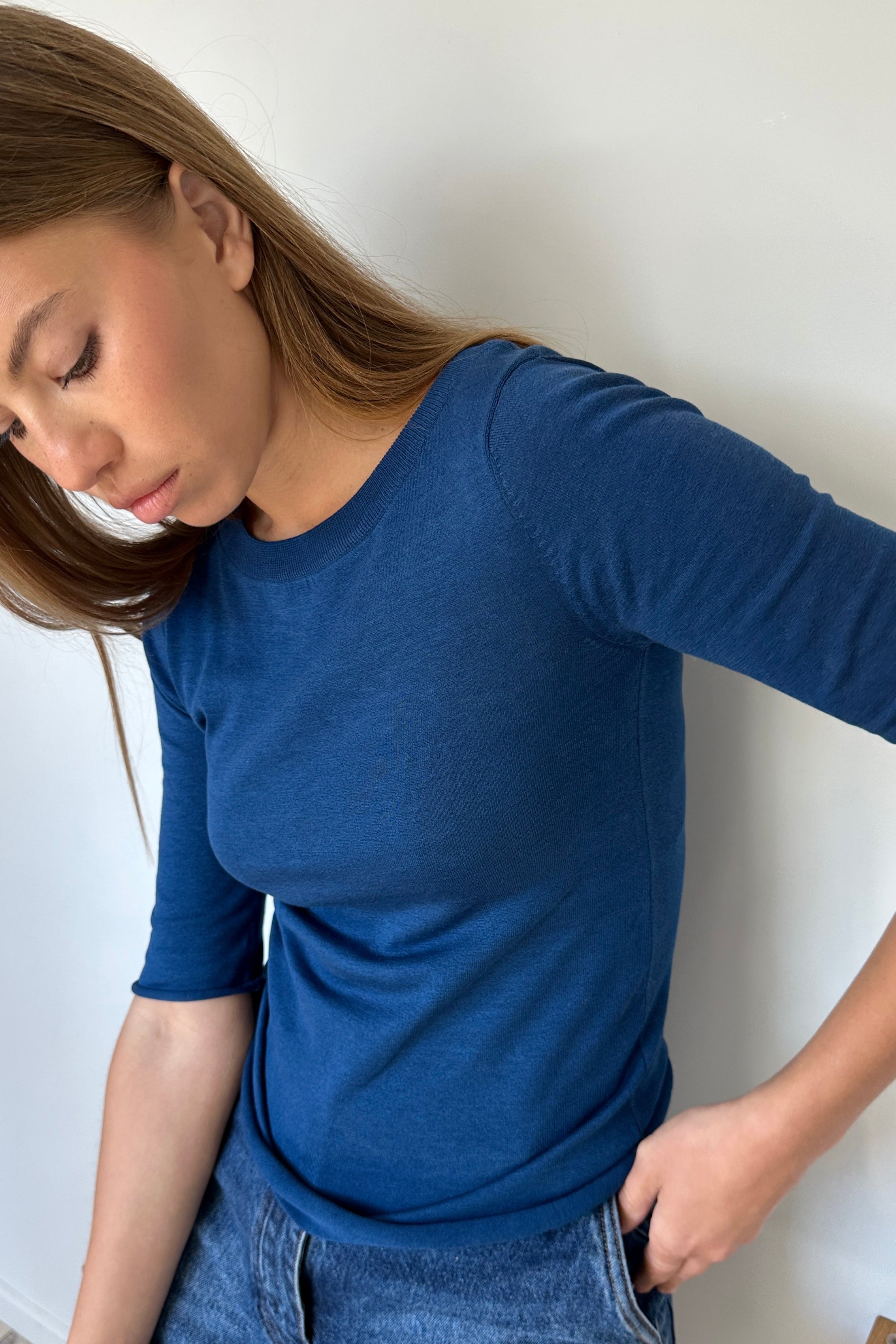 Woman wearing a blue long-sleeve shirt and jeans against a white wall.