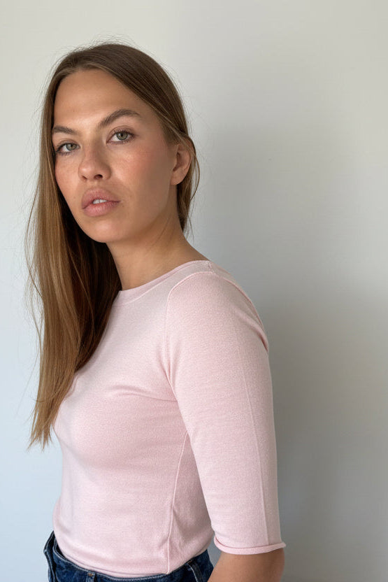 A woman wearing a fitted short-sleeve pale pink top and high-waisted blue jeans, standing indoors against a plain white wall.