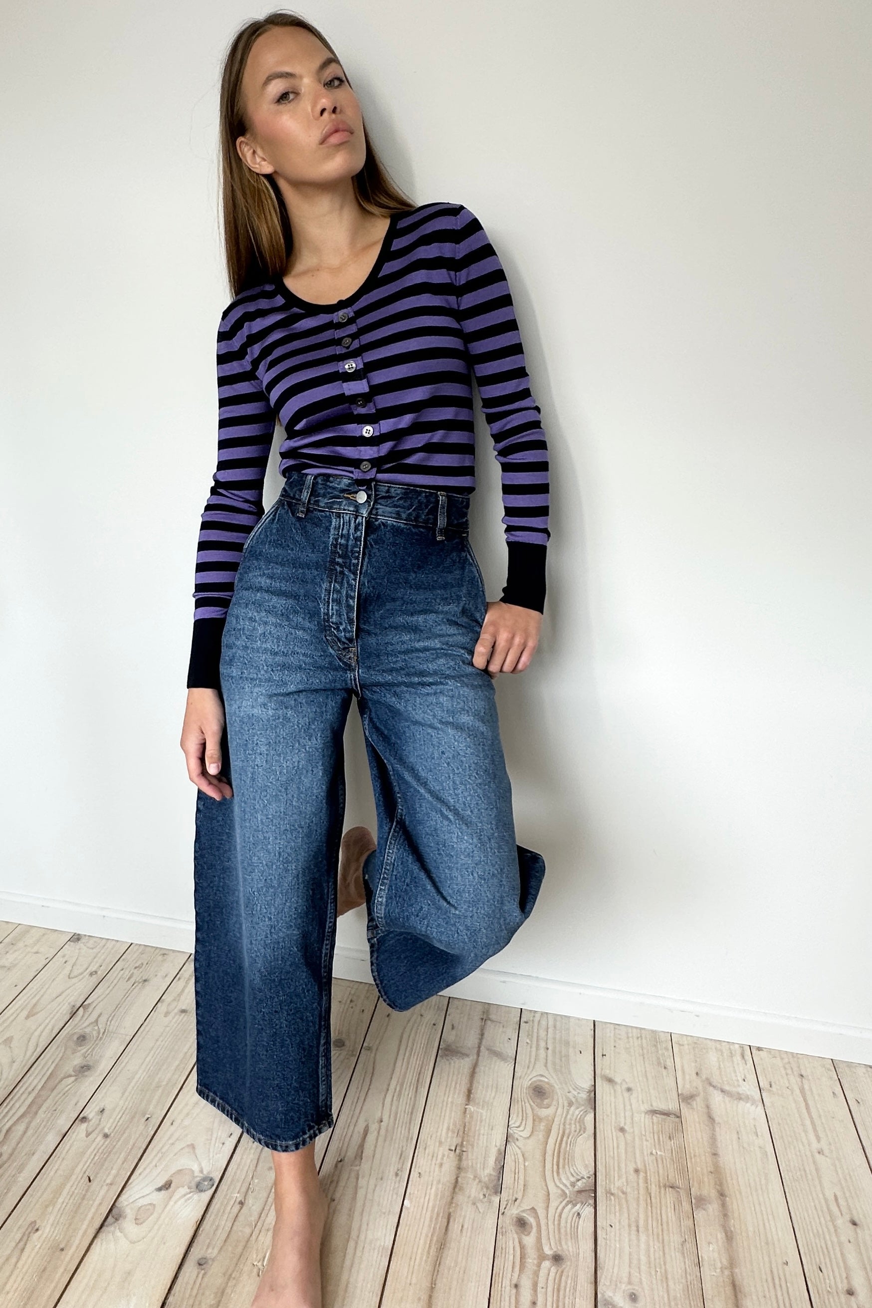 Person wearing a striped shirt and blue jeans against a white wall.