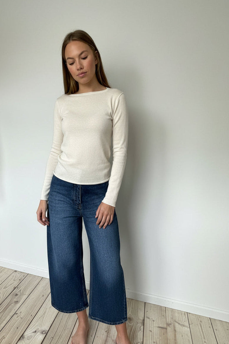 Woman wearing an ivory sweater and blue jeans standing against a white wall.