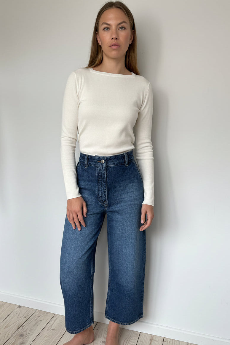 Person wearing an ivory long-sleeve top and blue jeans against a white wall.