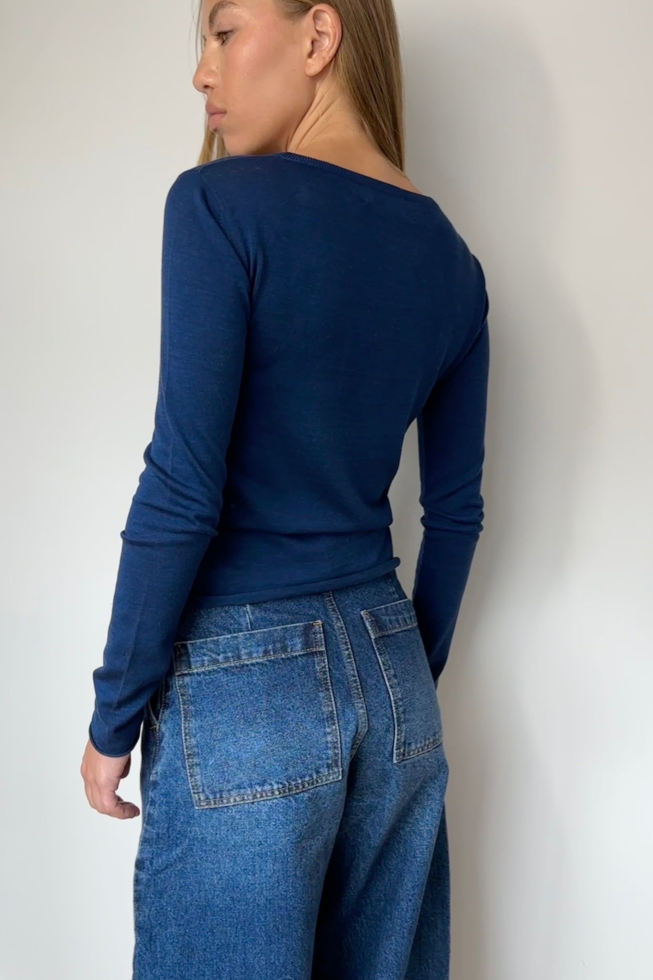 Person wearing a blue long-sleeve top and jeans against a white wall.