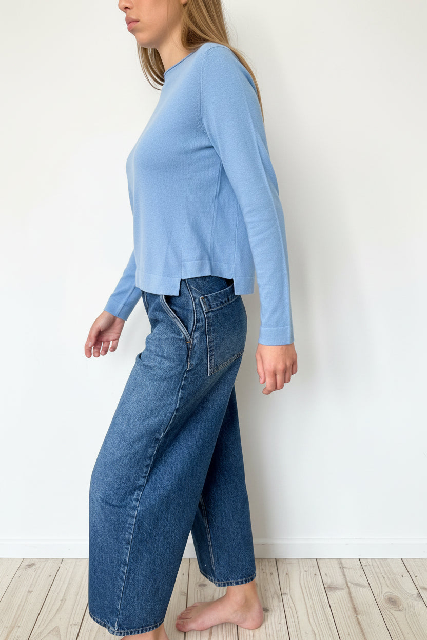 Person wearing a green shirt and blue jeans standing against a white wall.