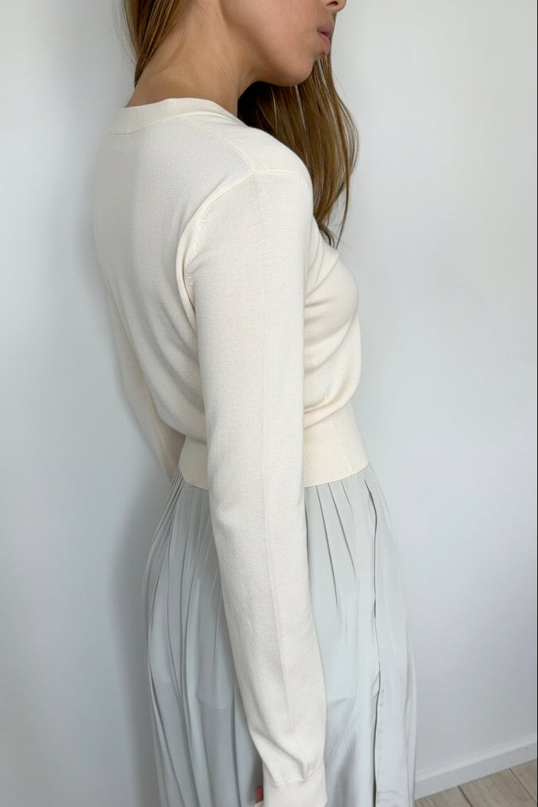 A woman shown from the side/back wearing an ivory long-sleeve knit top and a light gray pleated skirt, standing against a plain white wall.
