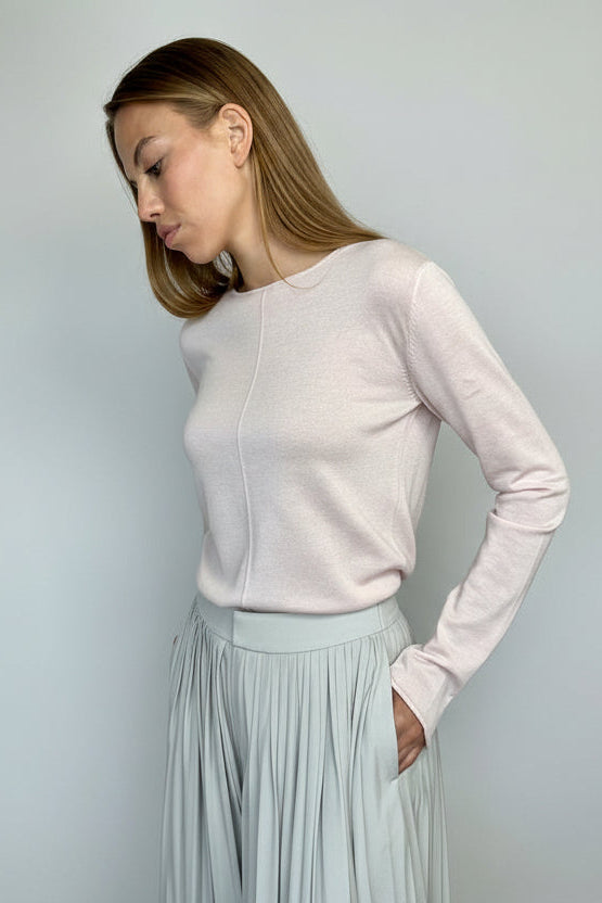 Woman wearing a light pink long-sleeve top and light gray pleated skirt against a plain background