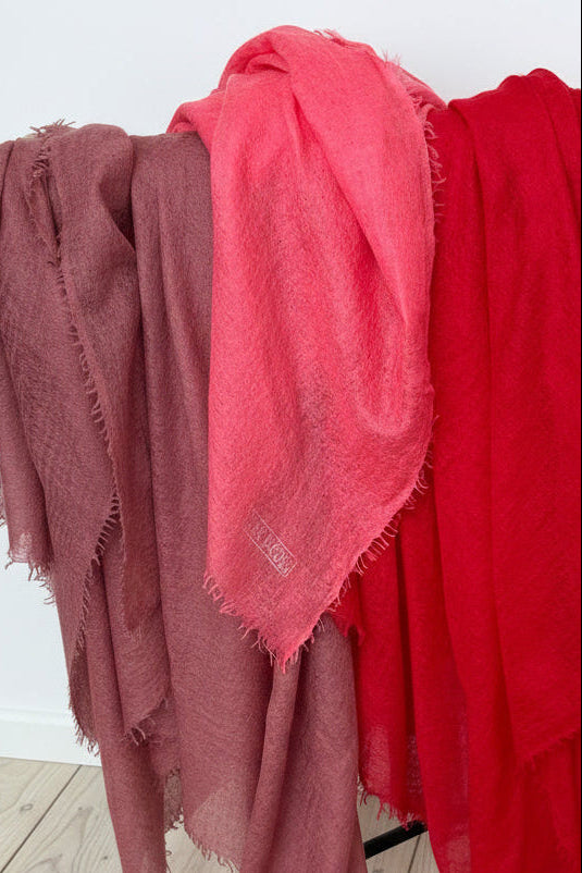 Three scarves in red, pink, and brown draped over a rack against a white wall.