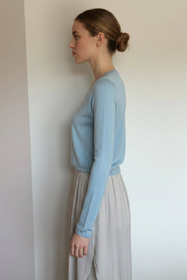 Woman wearing a light blue sweater and beige skirt against a plain wall.