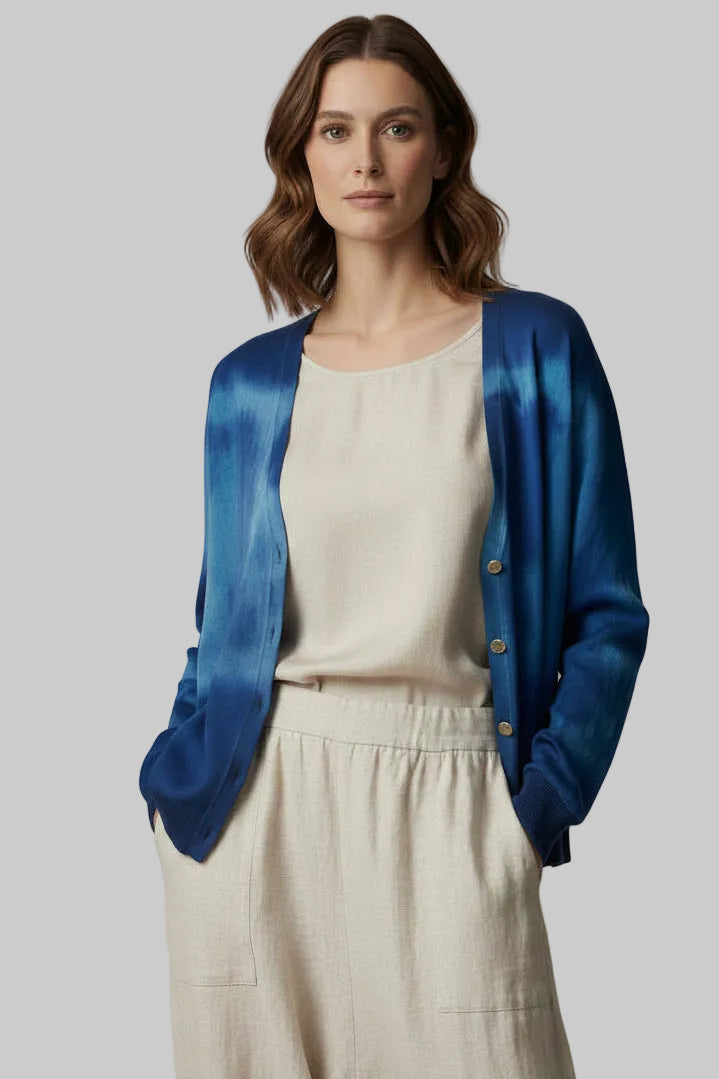 A woman modeling a vibrant blue tie-dye cardigan over a cream-colored top and matching pants, set against a plain light gray studio background.