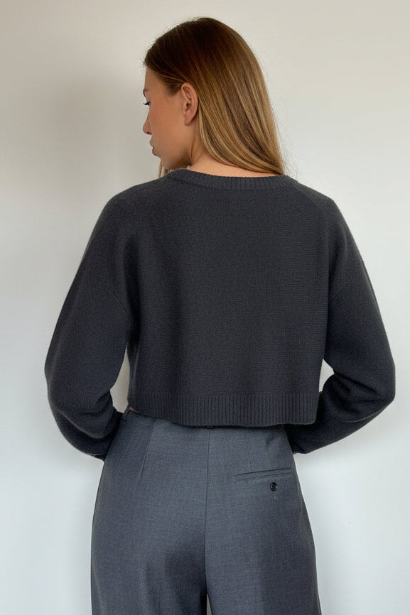 A woman wearing a dark charcoal gray cropped long-sleeve sweater and matching gray wide-leg trousers, photographed from the back against a plain off-white wall.