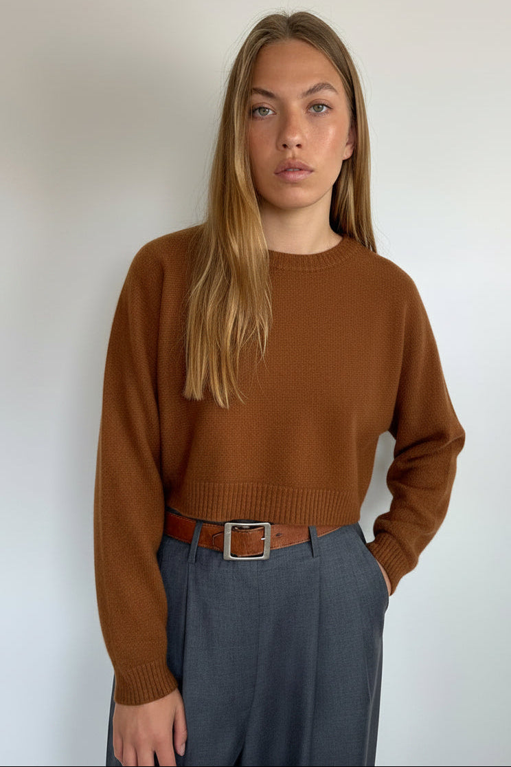 A woman modeling a dark charcoal gray knit sweater and matching gray trousers with a brown belt, standing against a plain off-white wall.
