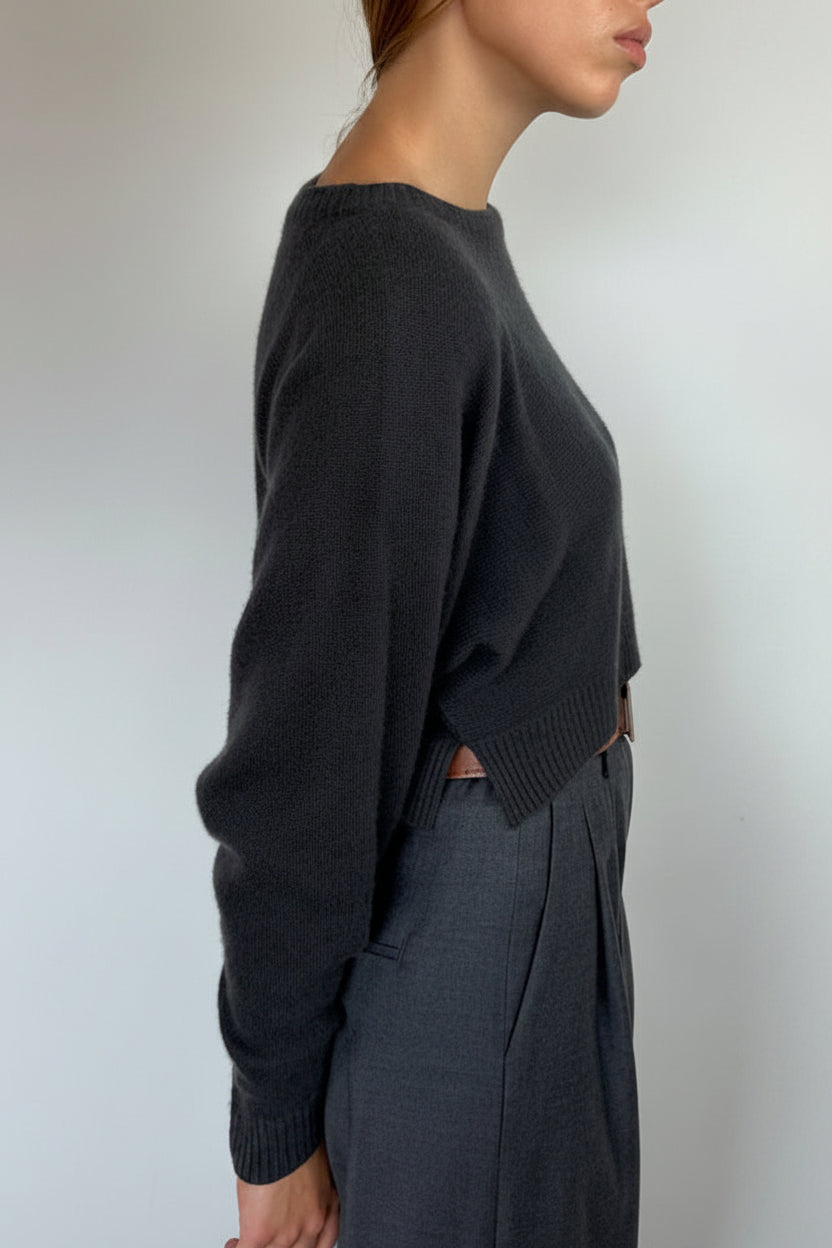 A woman shown in profile wearing a dark charcoal knit sweater and dark gray trousers against a plain light gray wall.