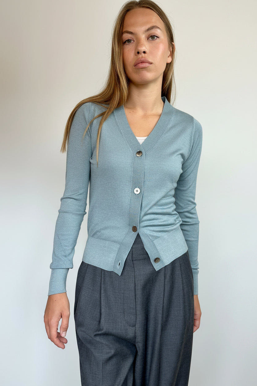 A woman modeling a light blue ribbed cardigan with a V-neck, three buttons, and a twisted hem detail, paired with dark gray wide-leg trousers, set against a plain off-white wall.