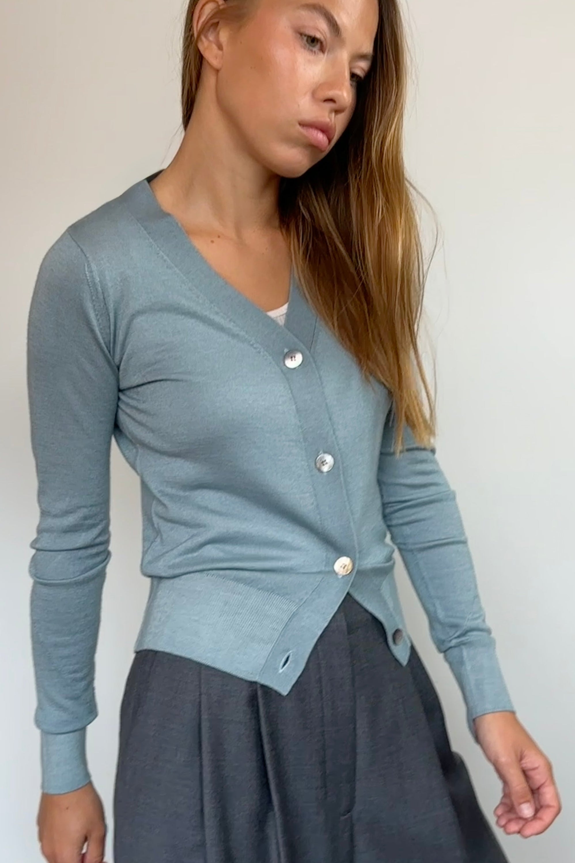 A woman wearing a light blue-gray long-sleeve cardigan with white buttons and a dark gray skirt, standing against a plain off-white wall.